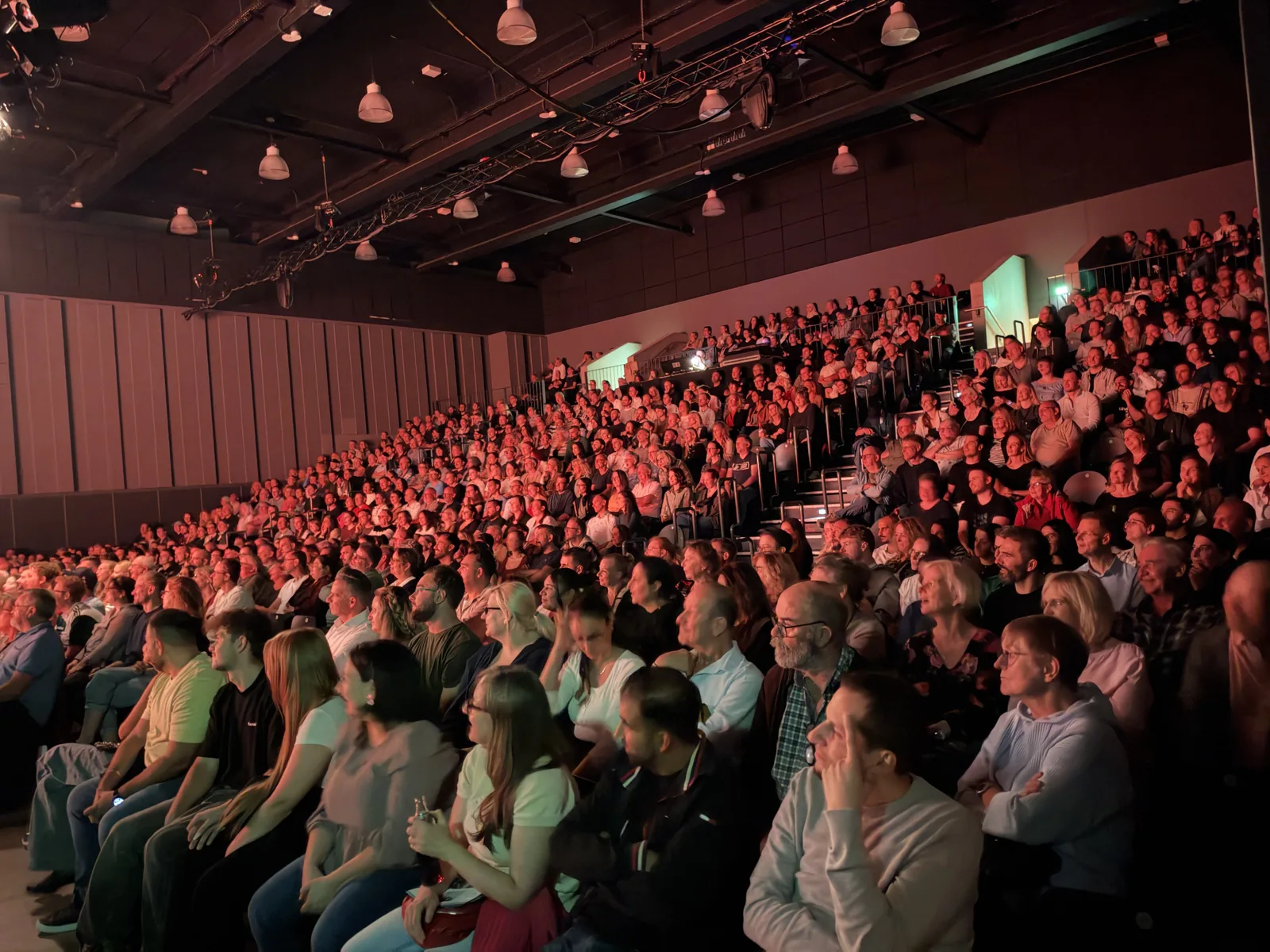 Ein voll besetzter Saal in ansteigenden Reihen, in warmes Bühnenlicht getaucht.